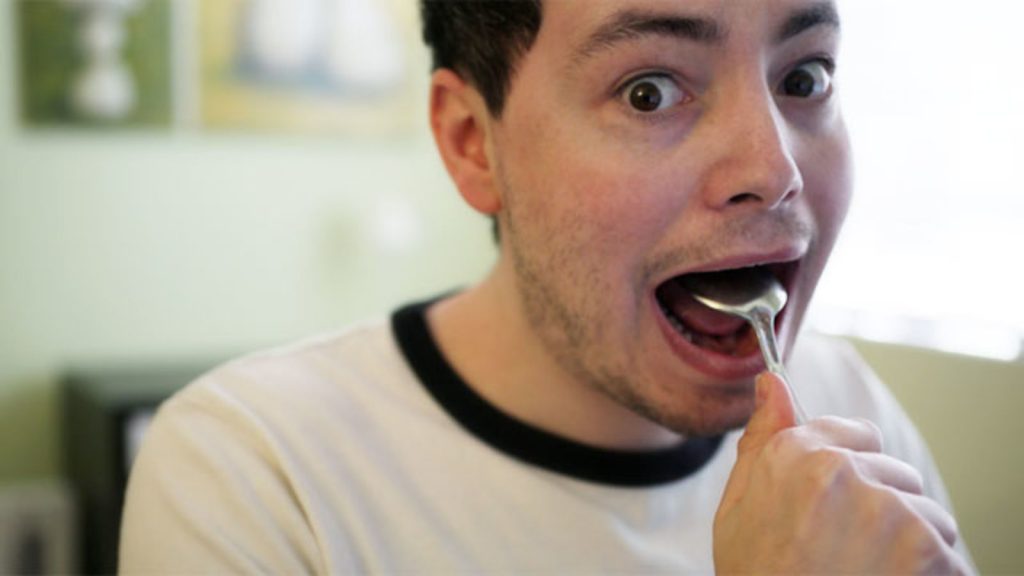 Image of man holding spoon on his tongue, inside of his mouth, with his eyes open wide.